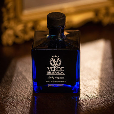 Bottiglia di olio extravergine d'oliva Verde Esmeralda Baby Organic, dal design elegante in vetro blu scuro con tappo nero. L'etichetta riporta il logo del brand e la dicitura 'Aceite de Oliva Virgen Extra - Producto de España'. Lo sfondo sfocato presenta una cornice dorata su una superficie in legno.