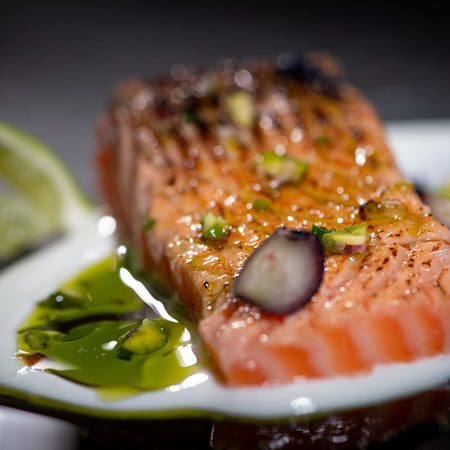 Primo piano di un filetto di salmone scottato, guarnito con erbe aromatiche, pistacchi tritati e cipolla rossa. Il piatto è decorato con crema bianca e gocce di olio extravergine d'oliva, che creano un effetto lucido e invitante. Accanto, una fetta di lime.