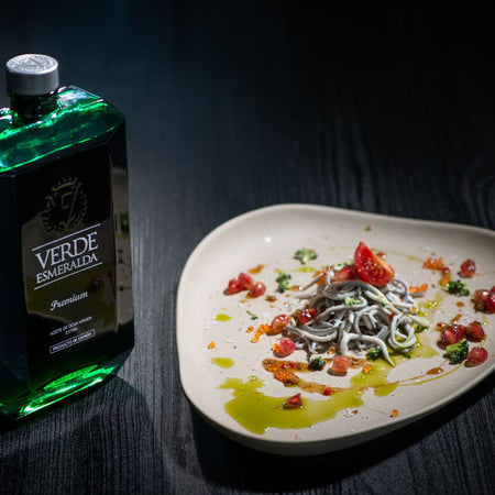 Piatto gourmet con angulas (piccole anguille) servite su un piatto dal design moderno, guarnite con pomodorini, melograno, prezzemolo e gocce di olio extravergine d'oliva. Sullo sfondo, bottiglia di olio Verde Esmeralda Premium in elegante confezione verde.