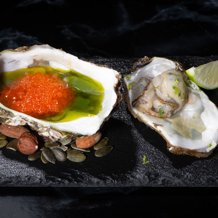 Piatto gourmet con due ostriche servite su un piatto di ardesia nera. Un'ostrica è guarnita con caviale rosso e olio extravergine d'oliva, mentre l'altra è al naturale con scorza di lime grattugiata. Accanto, semi di zucca, mandorle e una fetta di lime.