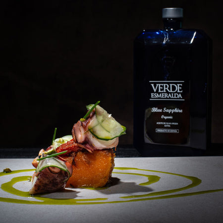 Piatto gourmet con polpo grigliato, fettine di zucchina e erba cipollina su una base croccante, decorato con gocce di olio d'oliva. Sullo sfondo, bottiglia di olio extravergine d'oliva Verde Esmeralda Blue Sapphire Organic, in un'elegante confezione blu.