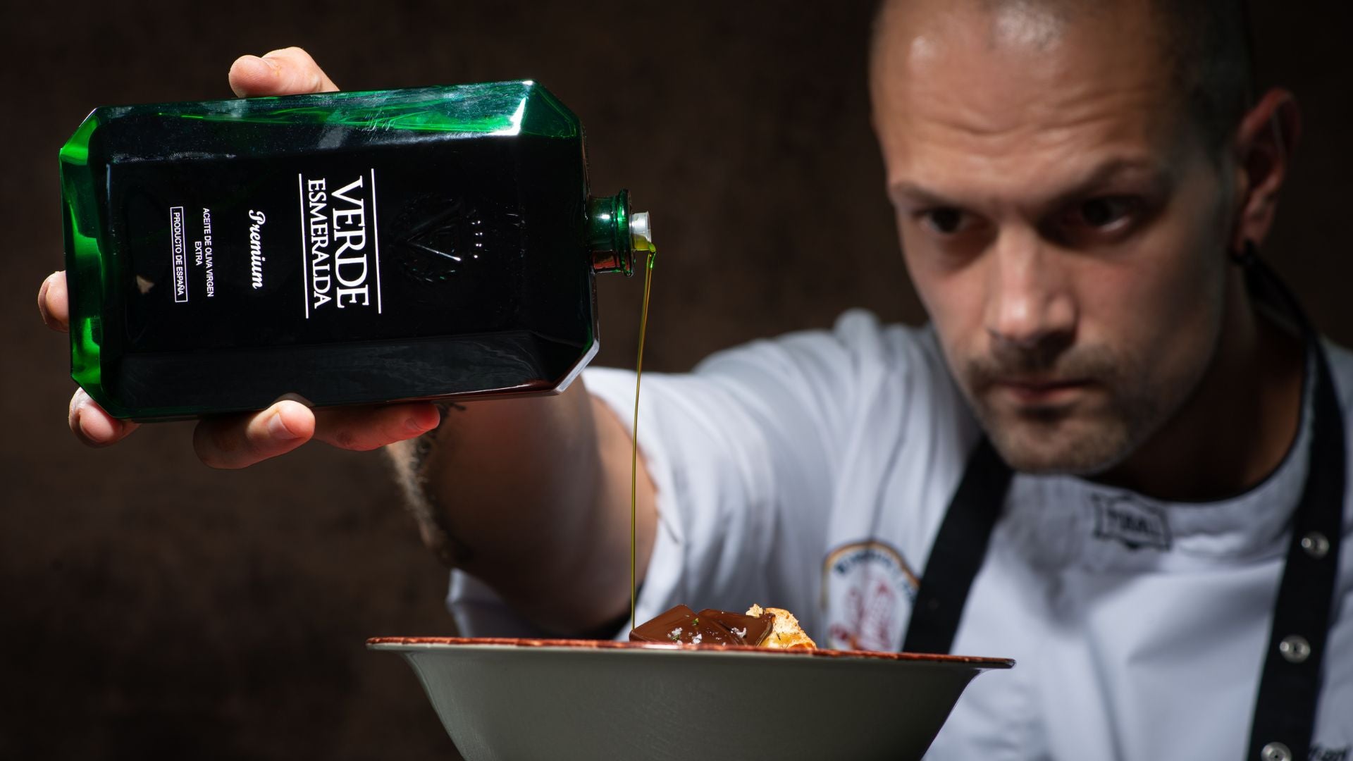 Uno chef concentrato versa con precisione olio extravergine d'oliva Verde Esmeralda da una bottiglia dal design elegante su un piatto gourmet. Il flusso dorato dell'olio enfatizza la qualità premium del prodotto, mentre lo sfondo scuro aggiunge un tocco di raffinatezza e lusso.